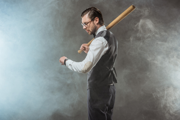 side view of stylish businessman holding baseball bat and checking wristwatch in smoke - Foto, afbeelding