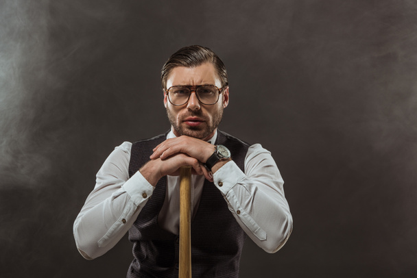 serious stylish man in eyeglasses leaning at baseball bat and looking at camera on black - Foto, Imagem
