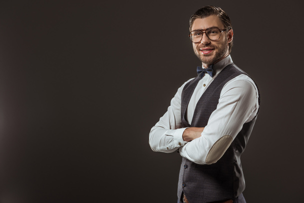 portrait of handsome stylish man in bow tie and eyeglasses standing with crossed arms and smiling at camera isolated on black - Photo, Image