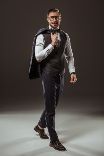 weergave van de volledige lengte van knappe stijlvolle man in bow tie en brillen houden pak jas en glimlachend in de camera op zwart - Foto, afbeelding