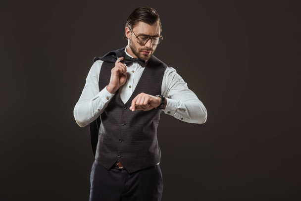 stylish businessman in bow tie and eyeglasses holding suit jacket and checking wristwatch isolated on black - Photo, Image