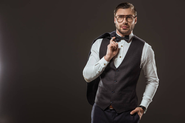 handsome stylish man in bow tie and eyeglasses holding suit jacket and looking at camera isolated on black - Foto, Imagem