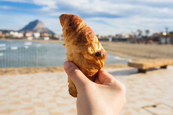 Croissant in der Hand vor dem Hintergrund eines schönen Strandes - Foto, Bild
