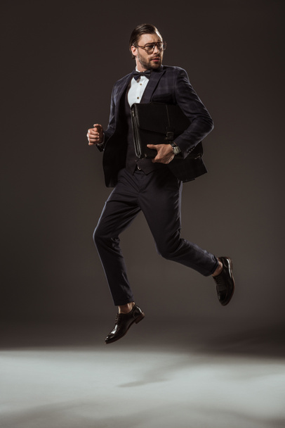 stylish businessman with briefcase jumping and looking away on black - 写真・画像