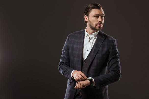 handsome stylish businessman checking wristwatch and looking away isolated on black - Photo, Image
