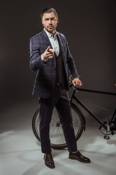 serious young man in suit pointing at camera while standing near bicycle on black  - Foto, immagini