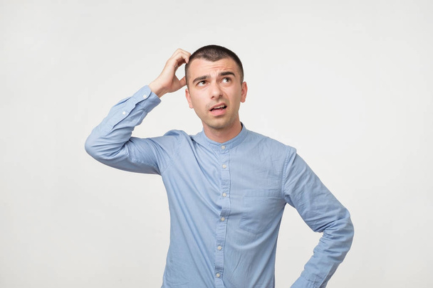 Handsome spanish guy in blue shirt, frowning and looking unsatisfied while scratching head, standing over gray background. He can not remember some facts or what to do. Problems with memory - Photo, Image