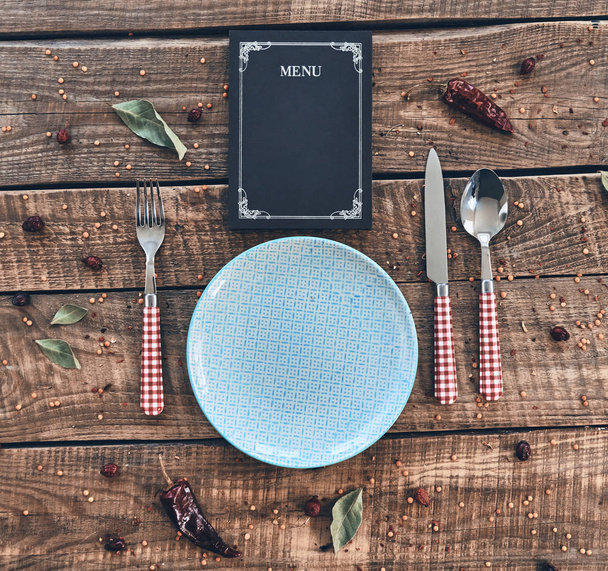 flat lay shot of empty plate, fork, spoon, knife and closed menu lying on wooden table - 写真・画像