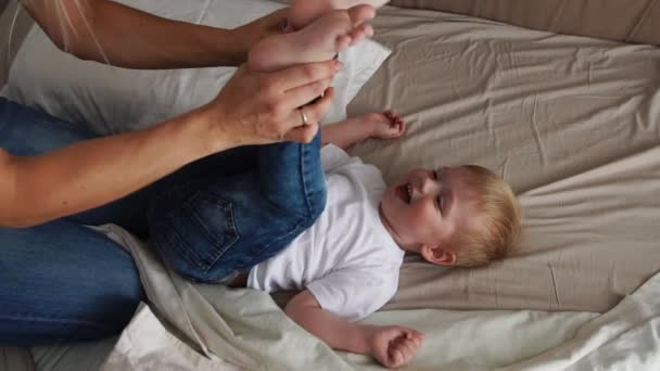 Mamá hace cosquillas al niño acostado en la cama que mira directamente a la cámara y se ríe en voz alta
 - Metraje, vídeo