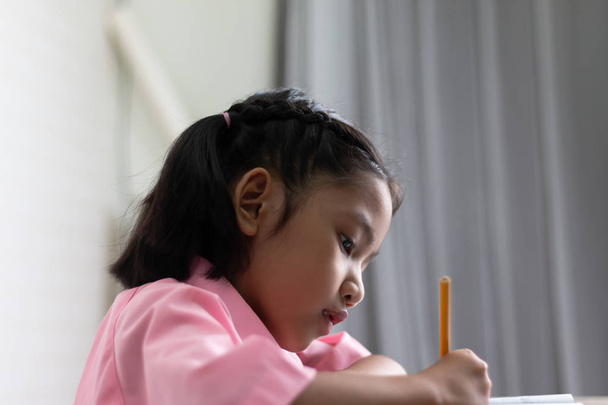 Close up the little girl is doing homework intently. Children use a yellow pencil is writing a notebook. Select focus shallow depth of field with copy space on white background. - Fotografie, Obrázek
