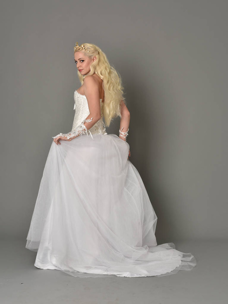 full length portrait of blonde girl wearing white gown, standing pose with back to the camera. grey studio background. - Photo, Image