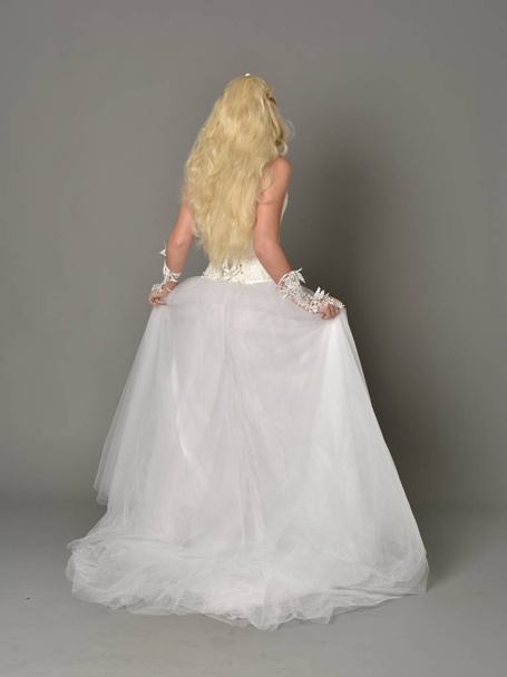full length portrait of blonde girl wearing white gown, standing pose with back to the camera. grey studio background. - Photo, Image