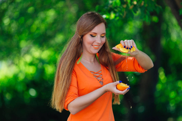 Porträt einer hübschen Frau mit rotem Haar, die im sommergrünen Park mit den Händen zwei Hälften saftig köstlicher Orangen presst. - Foto, Bild