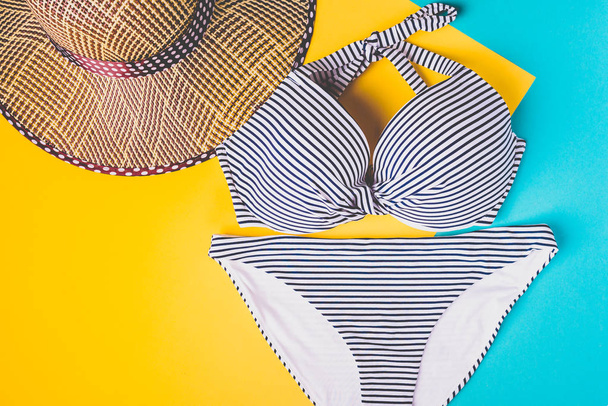 Top view of two pieces striped blue and white swimming suit and straw beach hat on blue and yellow pastel background. Concept of vacation. - Photo, Image
