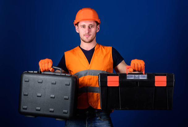 Man in helmet, hard hat holds toolbox and suitcase with tools, blue background. Toolbox and equipment concept. Worker, repairer, repairman, builder on thoughtful face choosing equipment for work - Photo, Image