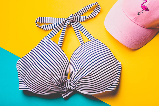 Вид сверху полосатого сине-белого купального бюстгальтера и розовой пляжной шляпы с фламинго на синем и желтом фоне. Принято. Концепция отпуска
. - Фото, изображение