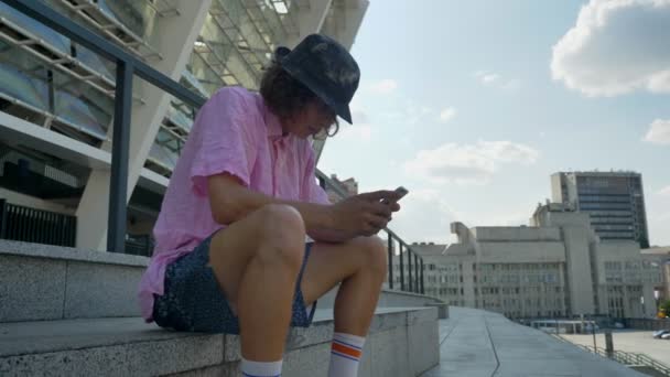 Young man hipster is typing message on smartphone, legs on skateboard, urban concept, communication concept, side view - Кадри, відео