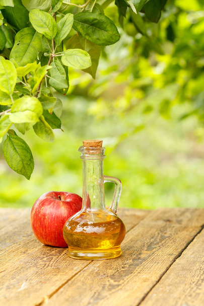 Яблочный уксус в стеклянной бутылке с пробкой и свежим красным яблоком на старых деревянных досках с размытым зеленым естественным фоном. Органическое питание для здоровья
 - Фото, изображение