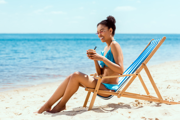 happy african american woman in bikini and sunglasses drinking cocktail in coconut shell while sitting on deck chair on sandy beach - Foto, imagen