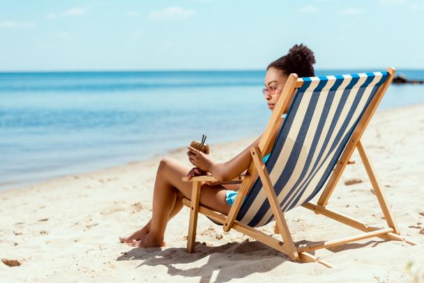 afrikai-amerikai nő a bikini és a gazdaság koktél a kókusz héj nyugágyat a tengerparton ülve napszemüveg  - Fotó, kép