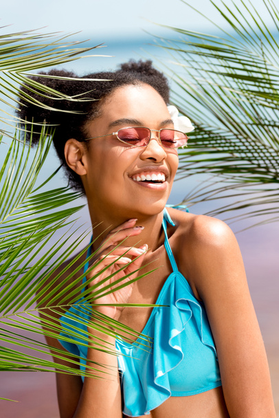 young smiling african american woman in sunglasses with flower in hair near palm leaves in front of sea - Photo, Image