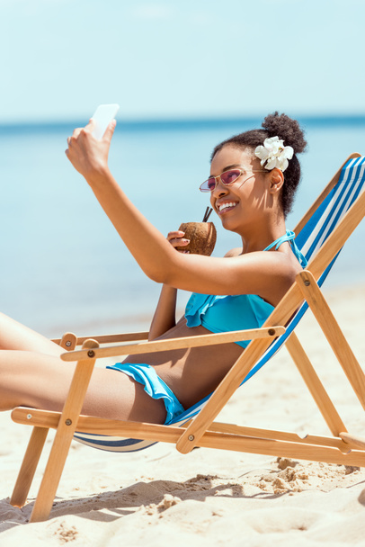 happy african american woman with flower in head drinking cocktail in coconut shell and taking selfie on smartphone while laying on deck chair on sandy beach  - Foto, Imagem