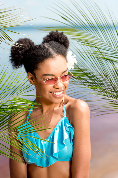 smiling african american woman in sunglasses with flower in hair near palm leaves in front of sea - Φωτογραφία, εικόνα