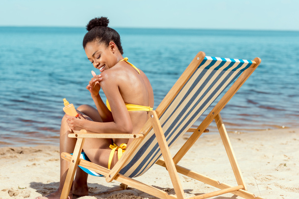 femme afro-américaine souriante appliquant de la crème solaire sur la peau tout en étant assis sur la chaise longue sur la plage de sable
  - Photo, image