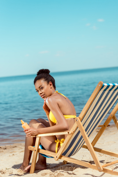 african american woman applying sunscreen lotion on skin while sitting on deck chair on sandy beach  - Fotoğraf, Görsel