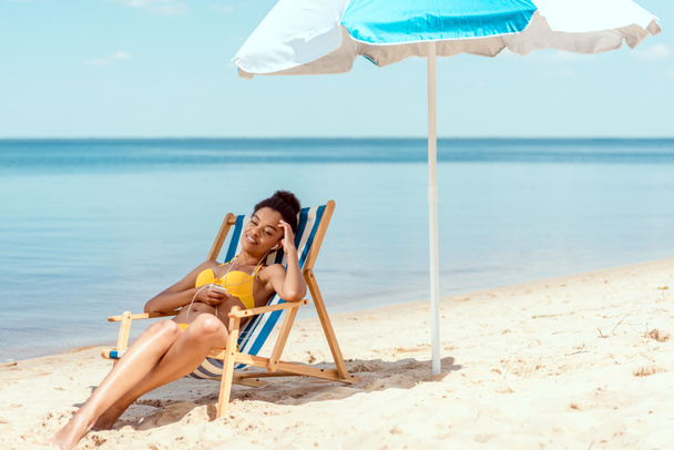 junge afrikanisch-amerikanische Frau entspannt sich im Liegestuhl und hört Musik in Kopfhörern mit Smartphone unter Sonnenschirm vor dem Meer  - Foto, Bild