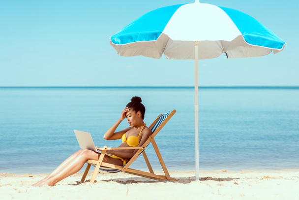 Zijaanzicht van lachende Afro-Amerikaanse vrouwelijke freelancer zittend op een ligstoel en met behulp van de laptop onder de parasol aan zee  - Foto, afbeelding