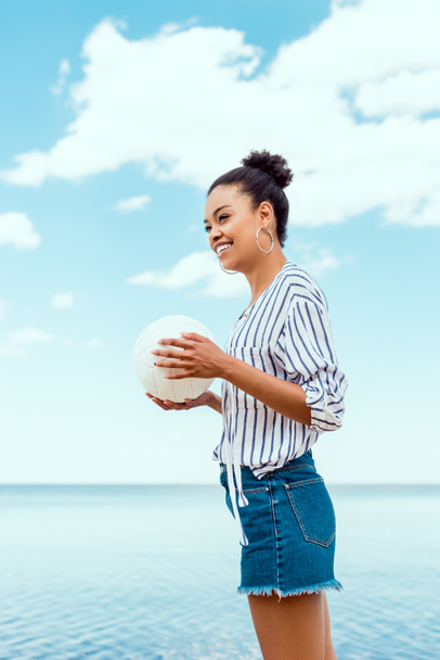vista basso angolo di donna afro-americana sorridente che tiene palla per pallavolo di fronte al mare
  - Foto, immagini