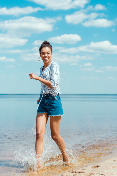onnellinen afrikkalainen amerikkalainen naisturisti kävelee merivedessä lähellä hiekkarantaa
 - Valokuva, kuva