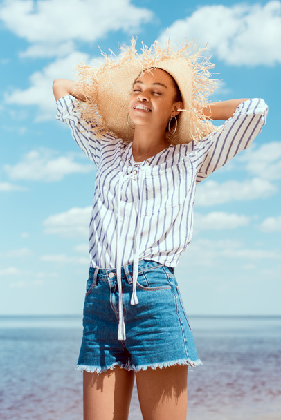 vista de ángulo bajo de viajero femenino afroamericano feliz en sombrero de paja frente al mar
  - Foto, Imagen