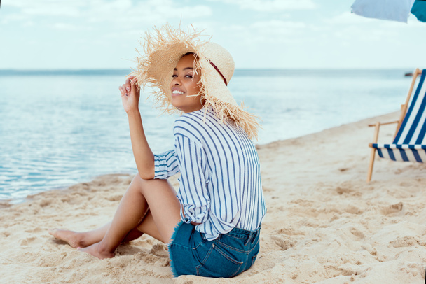 usmívající se africká americká žena v slamák, zatímco sedí na písečné pláži s lehátkem a slunečníkem při pohledu na fotoaparát  - Fotografie, Obrázek