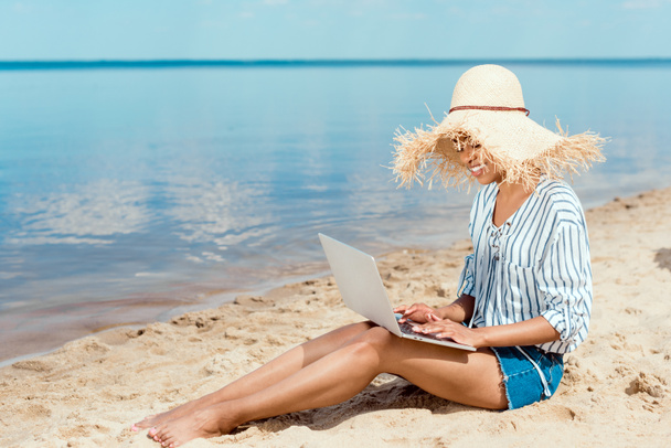feliz afroamericano freelancer femenino en sombrero de paja sentado en la playa de arena con el ordenador portátil
  - Foto, imagen