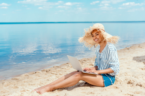 souriant afro-américaine femme pigiste en chapeau de paille assis sur la plage de sable avec ordinateur portable
  - Photo, image