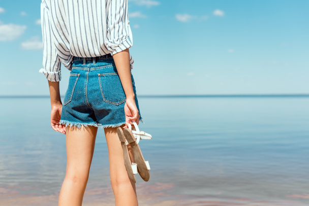 cropped image of young woman holding sandals in front of sea  - Фото, изображение