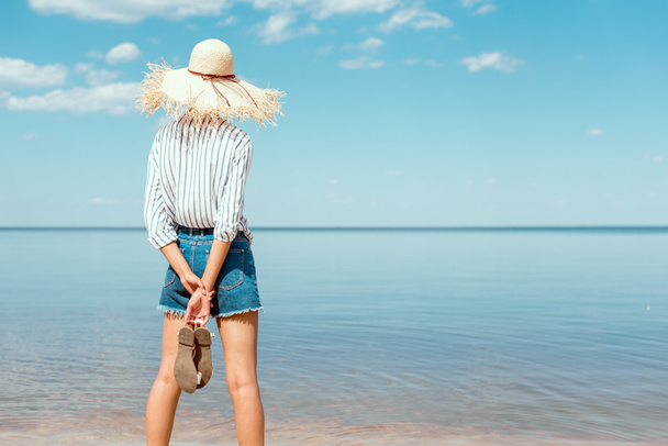 rear view of woman in straw hat holding sandals and looking at sea  - Foto, Bild