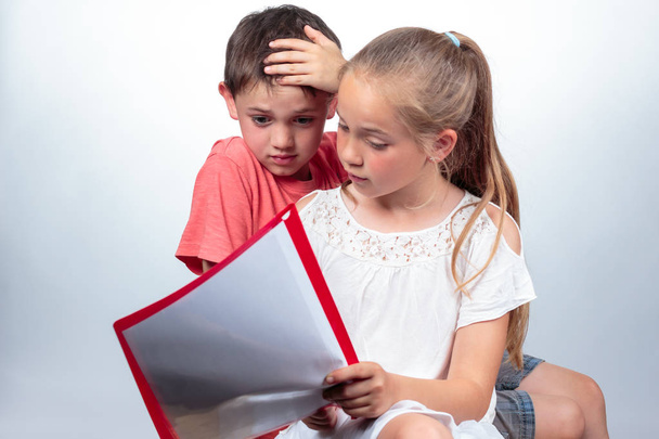Kaukasische Schulkinder lernen, ein nettes Mädchen hält Zettel in der Hand, erklärt ein Junge, der verwirrt aussieht und vor hellem Hintergrund seine Hand in die Haare schlägt. Bildungskonzept - Foto, Bild