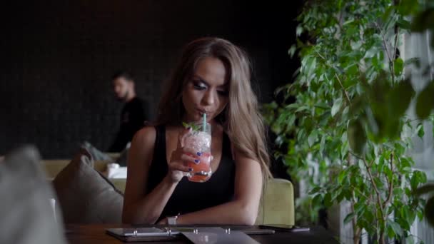 Cute girl is sitting in a cafe and having a cold drink indoor. Beautiful woman is enjoying hot summer day in cafe. Brushes her hair back. - Filmmaterial, Video