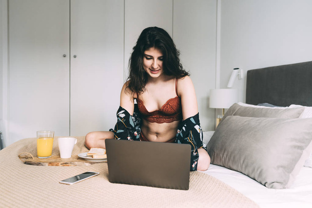 Ragazza in biancheria intima a fare colazione e utilizzando il computer portatile a letto
 - Foto, immagini