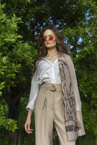 Beautiful stylish young girl wearing a trendy white shirt, beige trousers, pareo and red sunglasses poses outdoors against a background of green leaves. Commercial and advertisement design. Copy space - Foto, Bild