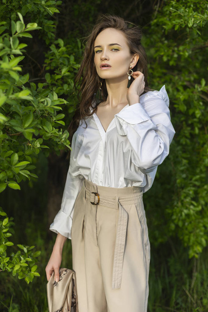 Beautiful stylish young girl wearing a trendy white shirt, beige trousers and a pareo poses outdoors against a background of green leaves. Nude makeup. Commercial and advertisement design. Copy space. - Foto, Imagen