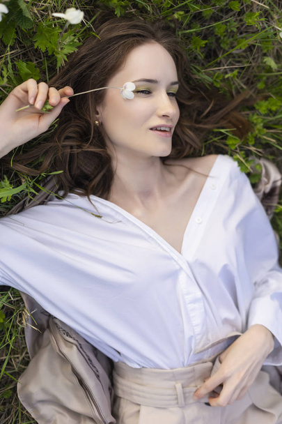 Beautiful stylish young girl wearing a trendy white shirt, beige bloomers, and a pareo lies relaxing on the green grass among the wild flowers. Commercial and advertisement design. Copy space. - Photo, Image