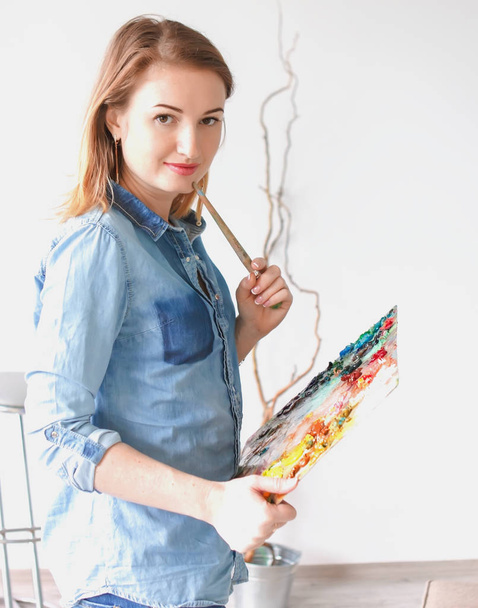girl artist with a palette of paints draws paintings in an art studio. Palettes and easel, creative mood - Fotoğraf, Görsel