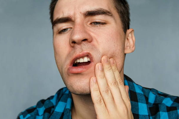 mladý muž s zub na modrém pozadí. Chlap s pohmožděnou čelist drží zadní části krku - Fotografie, Obrázek
