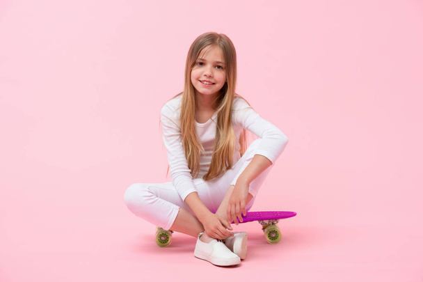 Happy child sit on penny board on pink background. Girl smile with skateboard. Little skater ready to ride. Skateboarding is fun. Active hobby and sport activity, punchy pastel trend. Childhood - 写真・画像