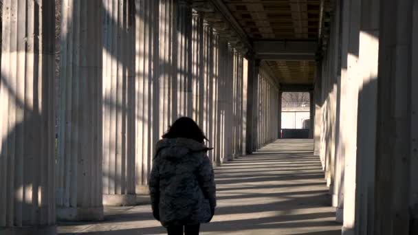 Rear view of young woman teenage girl mixed race female wearing camouflage jacket, walking through columns passageway during the day - Footage, Video