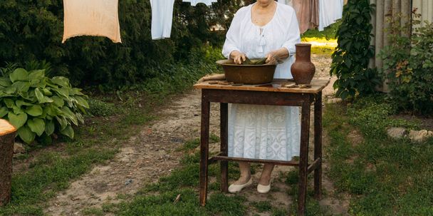 An elderly woman is washing clothes in the garden. in white vintage clothing. country life. clothes dries on a rope. next to the table are clothespins. Stark in an old copper basin and a clay jug. - Foto, Imagem